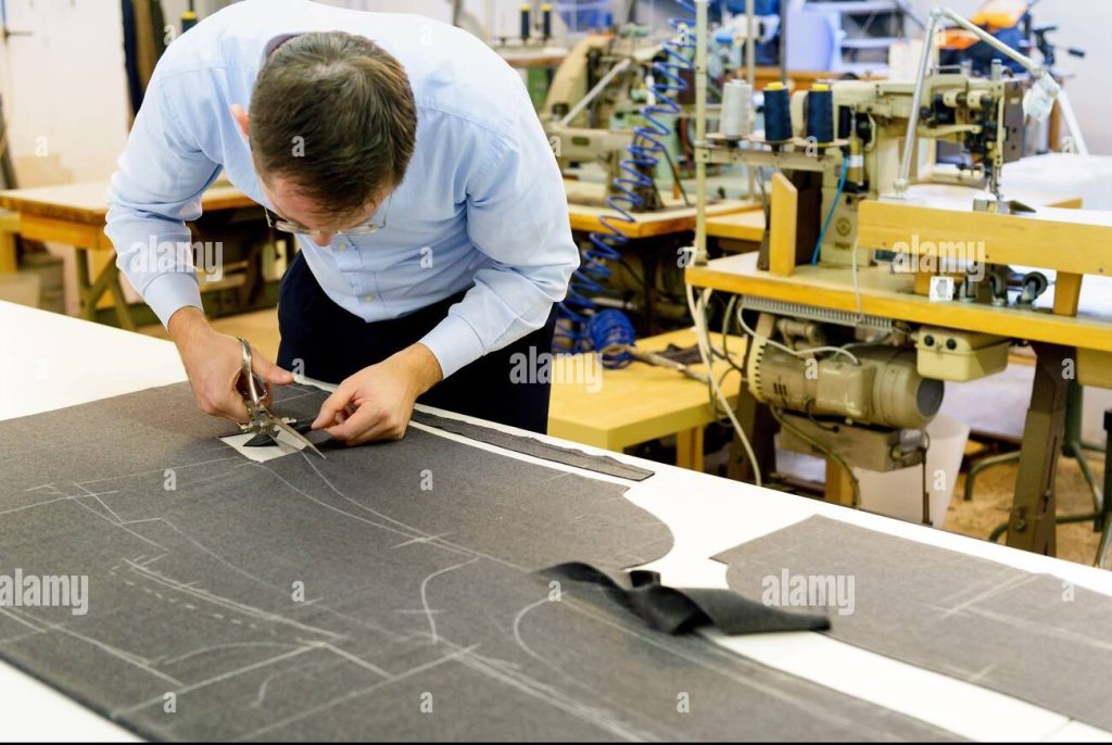 tailor bending over a workbench cutting fabric on which a pattern has been stencilled in a large spacious industrial workshop 2btd5w7